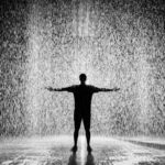 A person joyfully standing in the rain with arms wide open in a dramatic black and white scene.